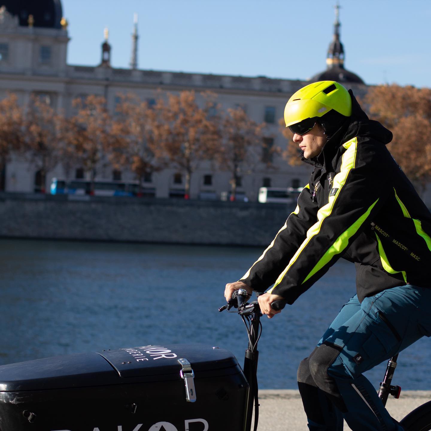 Rémi en livraison sur le vélo cargo Rakor, dôme de l'Hôtel-Dieu en arrière-plan, quais du Rhône, Lyon.